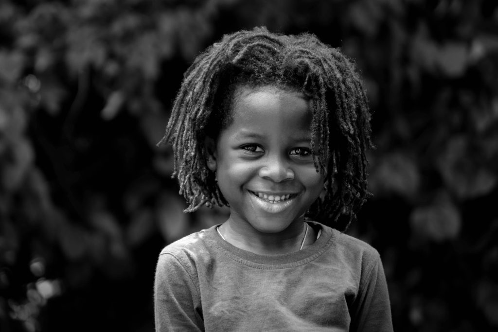 Adorable smiling child with dreadlocks posing outdoors, captured in black and white.